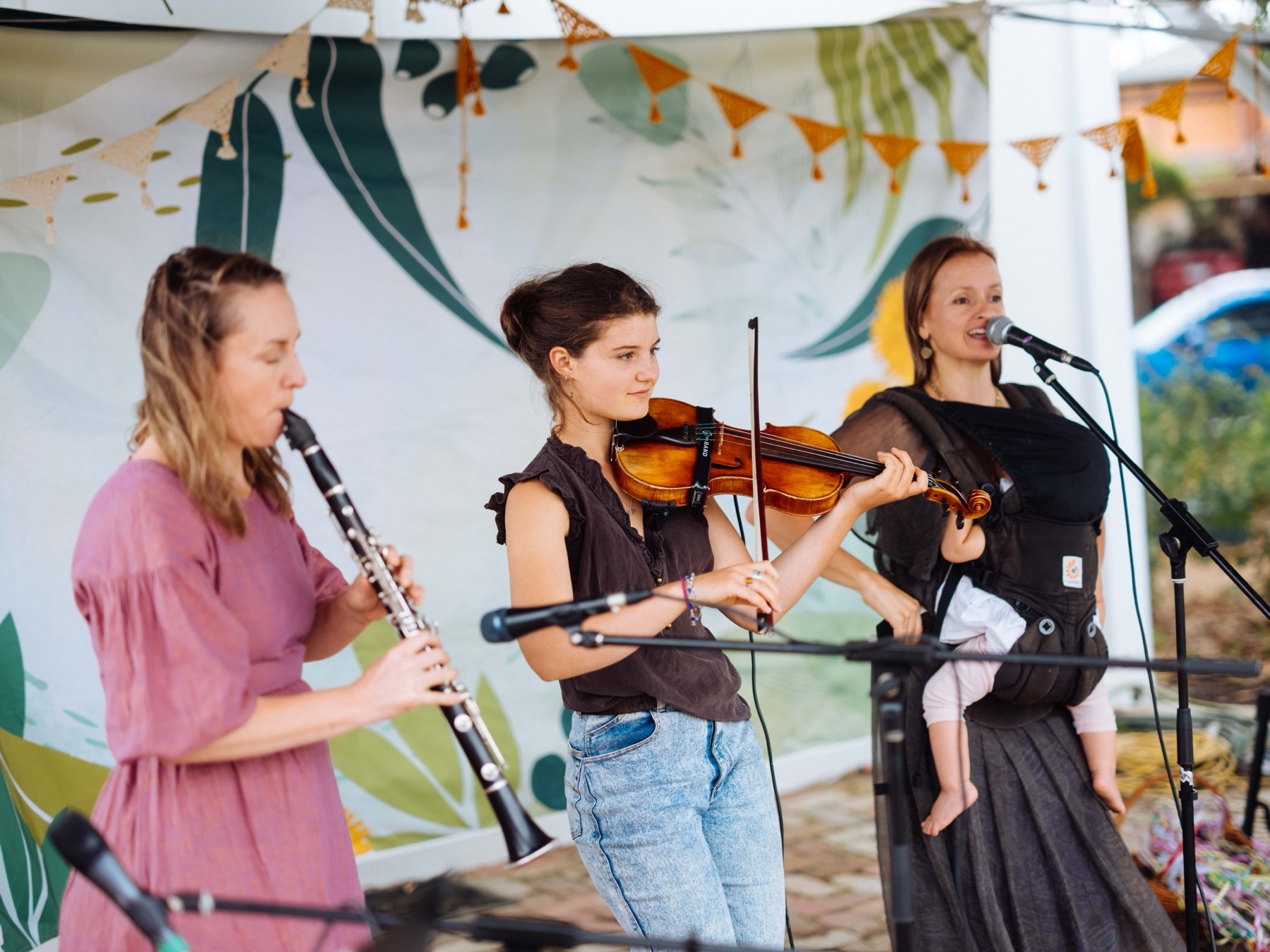 Rachel singing with a baby in a carrier beside a violin player and a clarinet player