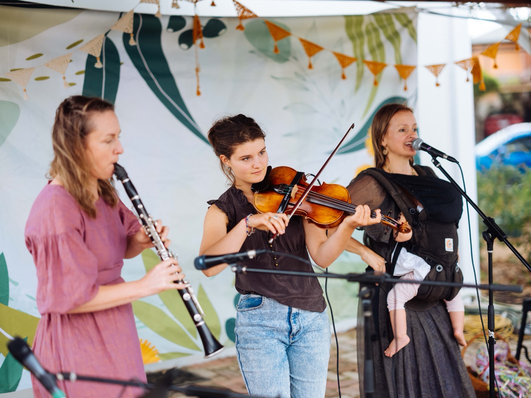 Rachel singing with a baby in a carrier beside a violin player and a clarinet player